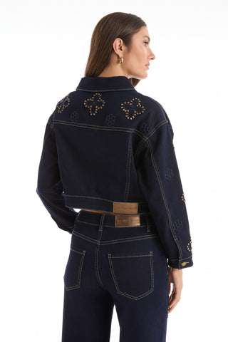 A woman faces away in the Kaia Studded Jacket - Indigo, featuring stud details and floral stitching, paired with matching high-waisted jeans. Her straight brown hair is loose, accessorized with gold hoop earrings, against a plain white background.