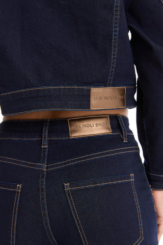 A close-up of a person wearing dark denim jeans and the Kaia Studded Jacket - Indigo, featuring brown leather patches reading "THE NOLI SHOP" near the waistband and back.