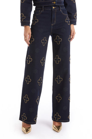 A person wears the Kaia Studded High Rise Jean in indigo with gold pointed-toe heels, standing against a white background.