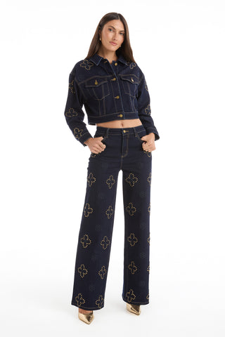 A woman stands confidently in the Kaia Studded Jacket - Indigo with matching high-waisted, wide-leg jeans featuring gold studs and floral stitching, paired with gold pointed-toe shoes against a white background.