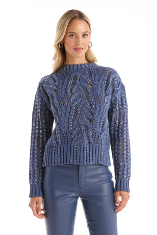 A blonde woman wears the Elea Crystal Knit Sweater in Sapphire and blue leather pants, standing against a plain white background.