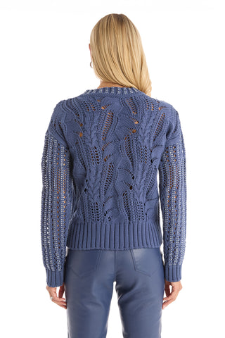 A woman with long blonde hair, seen from the back, wears the Elea Crystal Knit Sweater in Sapphire paired with blue leather pants against a white background.