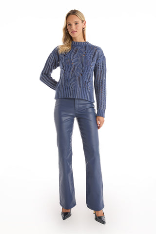 A woman with blonde hair and a neutral expression stands against a white background wearing the Elea Crystal Knit Sweater in Sapphire, high-waisted blue leather pants, and black pointed-toe heels.