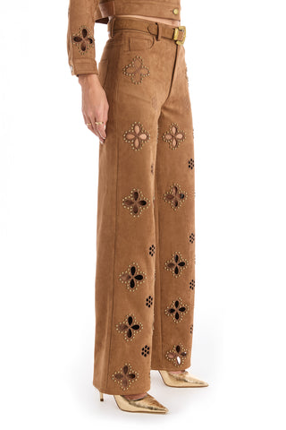 Kaia Jean wears the Kaia Suede High Rise Jean in Cognac—straight-leg stretch suede pants with large cut-out flower patterns, metal stud accents, a matching belt, and metallic gold pointed-toe heels against a white background.