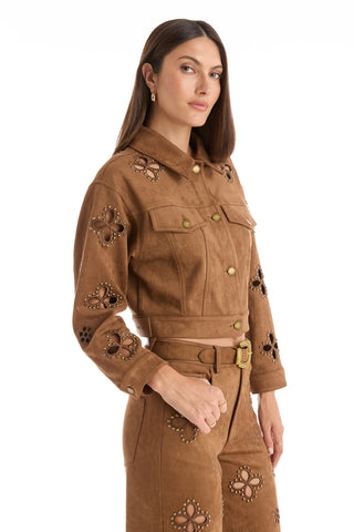 A woman with long brown hair models the Kaia Suede Jacket in Cognac, featuring decorative cutouts and stud details, paired with matching pants, standing against a white background.