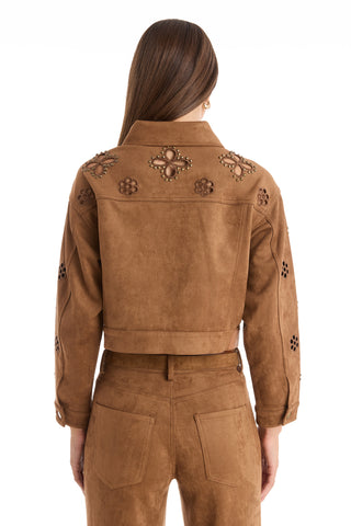 A woman faces away, wearing the Kaia Suede Jacket in Cognac with matching pants. The jacket features floral cutouts, decorative studs, and floral stitching on the back and sleeves. Her straight hair is worn down.