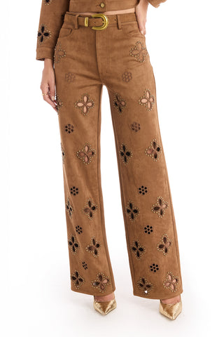 A person wearing the Kaia Suede High Rise Jean in cognac with decorative cutouts, a matching belt, and gold pointed-toe heels. Only the lower half of the outfit is visible.