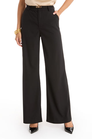 A person wearing Martina Belted Trouser - Black with a tucked-in beige top, black pointed-toe heels, and gold bangles stands against a white background.