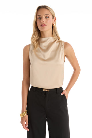 A woman with long blonde hair wears the Mara Silky Top in champagne, a sleeveless mock neck style, paired with black high-waisted pants. She accessorizes with gold hoop earrings and bracelets, standing confidently with one hand in her pocket.