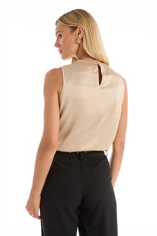 A woman with long blonde hair models the Mara Silky Top in Champagne, featuring a sleeveless mock neck and buttoned keyhole back, paired with black pants as she looks over her shoulder against a white background.
