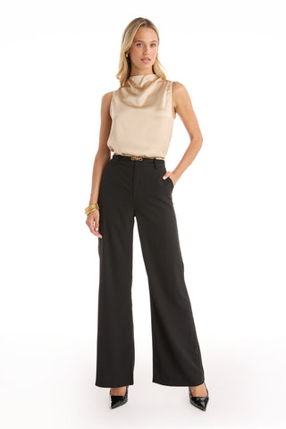 A woman poses against a white background with one hand in her pocket, wearing the Mara Silky Top in Champagne, high-waisted black wide-leg pants, black heels, and gold bracelets.