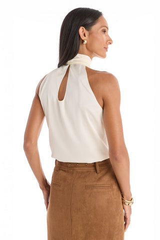 A woman with long dark hair models the Rina Tie Neck Blouse in Pearl, an elegant sleeveless top with a high neck and keyhole back, paired with a tan skirt. She faces away, head turned to the side, wearing hoop earrings and bracelets.