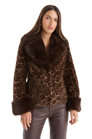 Wearing the Sydney Fur Trim Coat - Leopard with wide lapels and cuffs, paired with dark brown leather pants, a woman stands against a white background. This coat adds a bold touch to her chic cold-weather look.