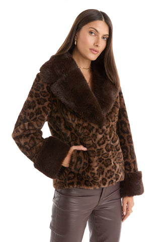 A woman wears the Sydney Fur Trim Coat - Leopard, featuring a wide collar and cuffs, paired with dark brown leather pants against a white background—a bold and chic choice for cold-weather style.