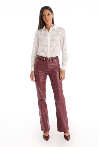 A woman with long dark hair stands facing forward, wearing a white blouse, burgundy leather pants, burgundy heels, and the Bettie Belt – Merlot with its pebbled faux leather finish and gold-toned buckle, set against a white background.