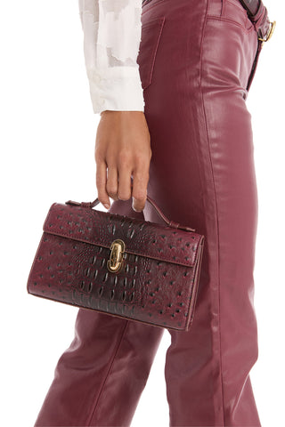 A person in high-waisted burgundy leather pants and a white long-sleeve shirt holds the Danica Ostrich Clutch - Merlot, a small textured burgundy bag with a gold clasp, by its top handle.