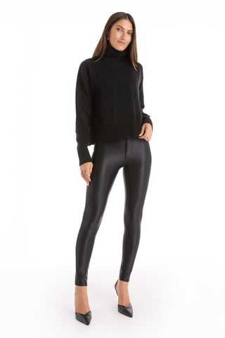 A woman stands against a white background wearing the Gia Ultra Soft Sweater in black, paired with shiny black leggings and pointed-toe heels. She has long straight hair and faces the camera with a neutral expression.