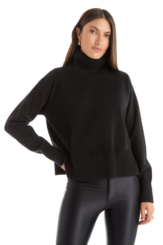 A woman with long brown hair models the Gia Ultra Soft Sweater in black, paired with fitted black pants and standing with one hand on her hip against a white background.