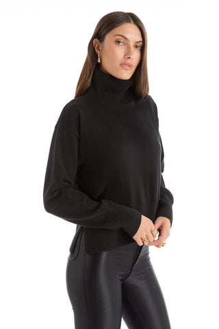 A woman with long brown hair models the Gia Ultra Soft Sweater in black with black leather pants, gold hoop earrings, and rings. She stands against a white background, facing slightly to the side with a neutral expression.