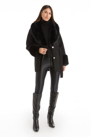 A woman faces forward in the Isabel Fur Trim Coat - Black, paired with a black turtleneck, shiny leggings, and tall boots, her straight hair and minimal jewelry enhancing the coat's elegance against a white background.