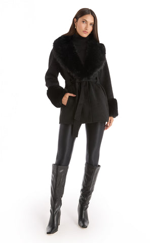 A woman stands against a white background wearing the Isabel Fur Trim Coat - Black, paired with a black turtleneck, shiny black leggings, and knee-high black boots.
