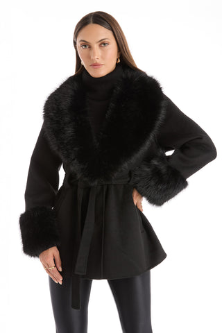 A woman wears the Isabel Fur Trim Coat - Black, paired with a black turtleneck sweater and black leggings, standing against a white background.