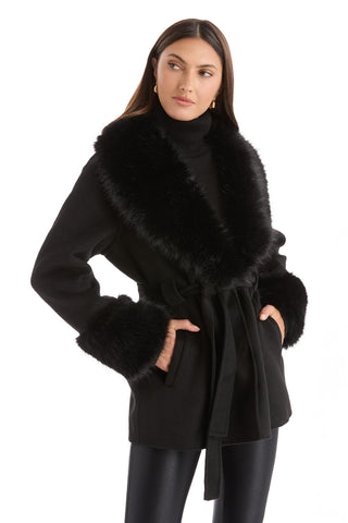 Woman wearing the Isabel Fur Trim Coat - Black with a draped silhouette, paired with a black turtleneck sweater and leather-look leggings, stands with one hand in her pocket, gazing to the side.