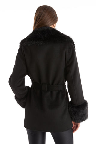 A woman with straight brown hair faces away, modeling the Isabel Fur Trim Coat - Black, featuring a belted waist and fur-trimmed collar and cuffs. She pairs this elegant coat with black leggings against a white background.