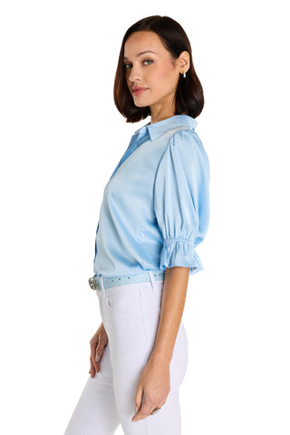 A woman with a short dark bob hairstyle models the Cindy Ruffle Sleeve Blouse in Sky Blue with high-waisted white pants, posing in profile against a plain white background.