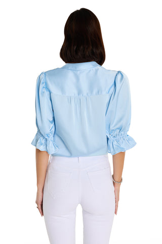 A woman stands with her back to the camera wearing the Cindy Ruffle Sleeve Blouse in Sky Blue, featuring ruffled cuffs, paired with high-waisted white pants against a plain white background.