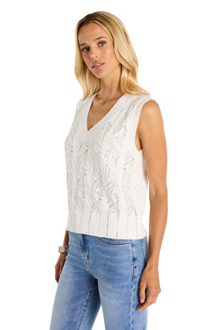 A woman with long blonde hair wears the Mikaela Crystal Knit Tank in white, featuring a textured V-neck design, paired with light blue jeans. She stands against a plain white background.