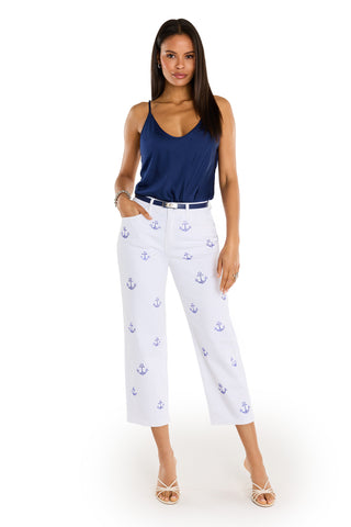 A woman faces forward, wearing the Kelsey Cropped Jean - Crystal Anchors featuring blue anchor patterns and a navy sleeveless top. She has one hand in her pocket, white heeled sandals on, and stands against a plain white background.