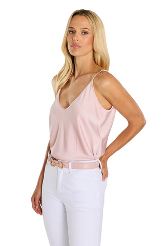A woman with long blonde hair poses against a white background in white pants and the Yara Silk Camisole - Blush, featuring adjustable straps.