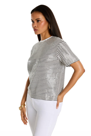 A woman with long dark hair and gold hoop earrings poses with one hand on her hip, wearing the Miranda Sequin T-Shirt in navy stripe and white pants against a plain white background.