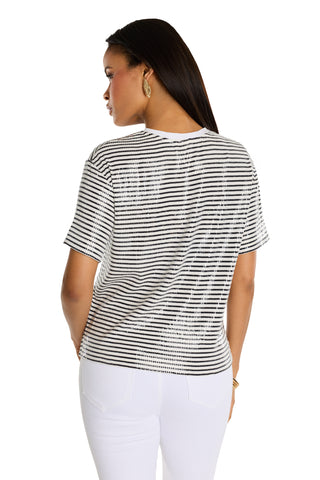 A woman with long dark hair stands facing away, wearing the Miranda Sequin T-Shirt - Navy Stripe and white pants, accessorized with gold hoop earrings and a bracelet against a plain white background.