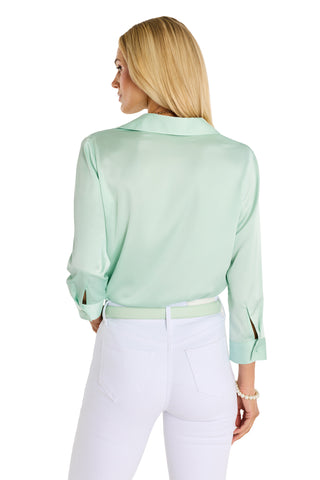 A woman with long blonde hair is seen from the back, wearing the Sara Silk Blouse in mint and white pants with a white belt. She creates a polished silhouette, resting one hand on her hip.