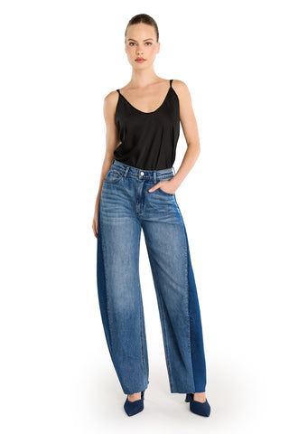 A woman stands against a white background wearing a black sleeveless top, Selena Barrel Jean - Medium Blue with wide legs and dual-tone denim side panels, and blue pointed-toe shoes. One hand rests in her pocket.