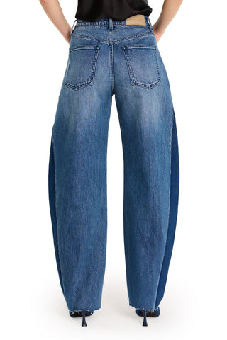 A person wears the Selena Barrel Jean - Medium Blue, high-waisted, wide-leg denim with exaggerated side panels and dual-tone design; hands on hips, showing only lower torso and legs against a white background.