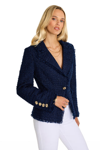 A woman with long blonde hair wears the Giselle Single Breasted Tweed Blazer in navy, featuring gold knot buttons and padded shoulders, paired with white pants. She stands against a plain white background, looking at the camera neutrally.