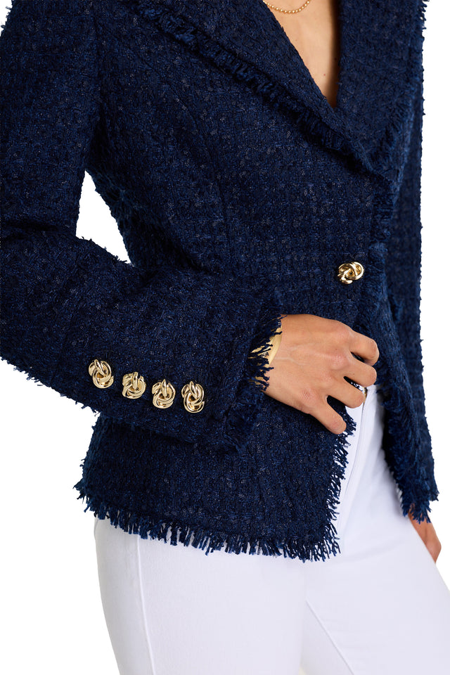 A person wearing the Giselle Single Breasted Tweed Blazer in navy, featuring gold knot buttons, frayed edges, padded shoulders, and paired with white pants. Their hand highlights a gold button and wristwatch near the waist.