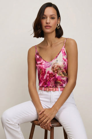 A woman with short brown hair sits on a wooden stool, wearing the Yara Silk Camisole in Bright Orchid, paired with white pants, a pink belt, and gold hoop earrings, posing against a plain light background.