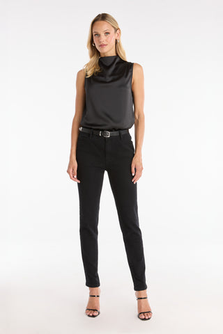 A woman with blonde hair stands against a white background, wearing the Mara Silky Top - Black, paired with black jeans, a belt, and black heeled sandals. She has a neutral expression.