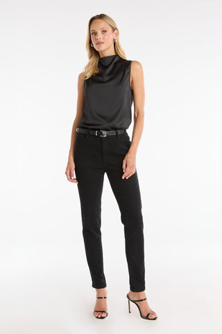 A woman with blonde hair and a neutral expression wears the Mara Silky Top in black, styled with black belted pants and black high-heeled sandals, standing against a white background.