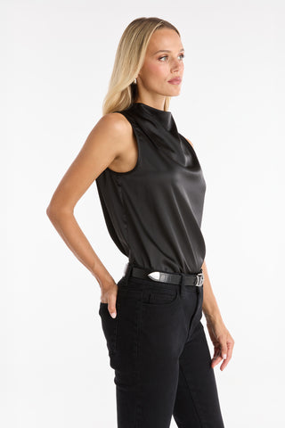 A woman with long blonde hair models the Mara Silky Top in black, a sleeveless mock neck satin style, paired with black jeans. She stands against a white background, one hand in her pocket, gazing to the side.