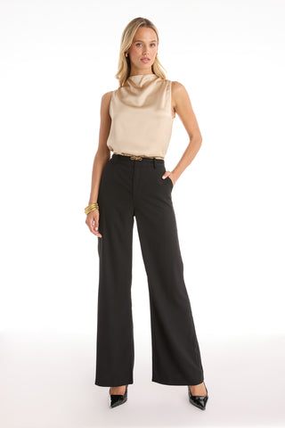 A woman stands confidently in the Mara Silky Top - Champagne, paired with black high-waisted wide-leg pants, black heels, and gold accessories, posing with one hand in her pocket against a plain white background.
