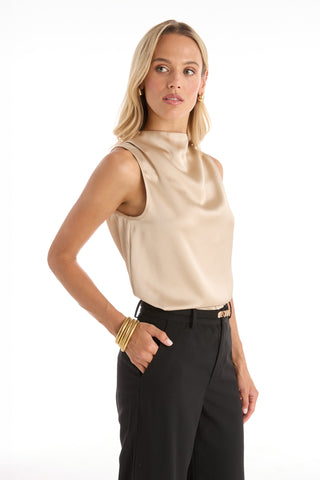 A woman with long blonde hair wears the Mara Silky Top in Champagne, paired with black trousers, gold hoop earrings, and bangles. She stands with one hand in her pocket, looking to the side against a white background.