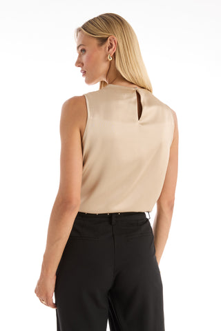 A woman with long blonde hair, wearing the Mara Silky Top in Champagne with a sleeveless mock neck and back keyhole detail, pairs it with black pants as she stands against a white background, slightly turning her head to the side.