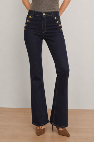 A person wearing the Allegra Bootcut Jean - Indigo, featuring a high-waisted, indigo wash silhouette with gold button details at the hips, styled with a fitted gray top and brown pointed-toe heels, standing on a wooden floor against a light wall.