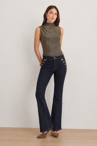 A woman wearing a sleeveless metallic top, brown heels, and the Allegra Bootcut Jean - Indigo with gold buttons stands against a light background.