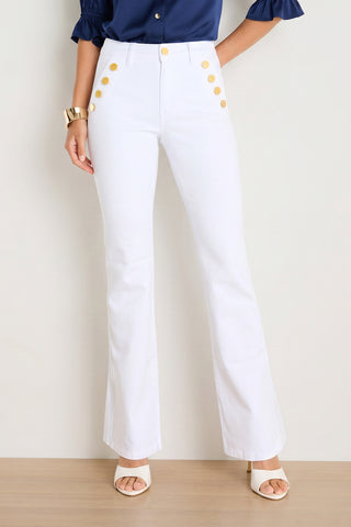 Wearing the Allegra Bootcut Jean in White—a chic, high-waisted stretch style with a flared silhouette and gold buttons—a woman pairs them with a navy blouse, white heeled sandals, and a gold bracelet, standing against a light background.